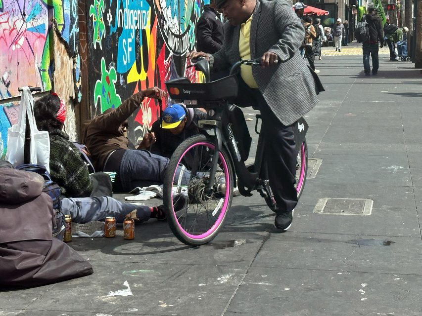 Three people sit on the sidewalk near a colorful graffiti wall, surrounded by bags and cans, as a person on a bike passes by.