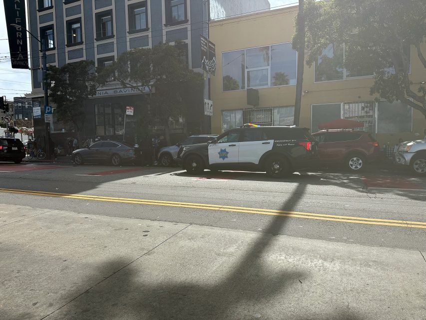 A police SUV is parked on a city street in front of a row of buildings on a sunny day.