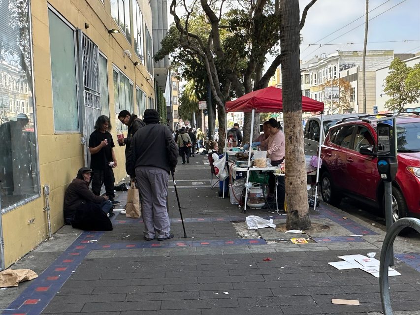 People gather on a city sidewalk with some sitting on the ground and others standing near a table under a red canopy selling food. Cars are parked along the street.