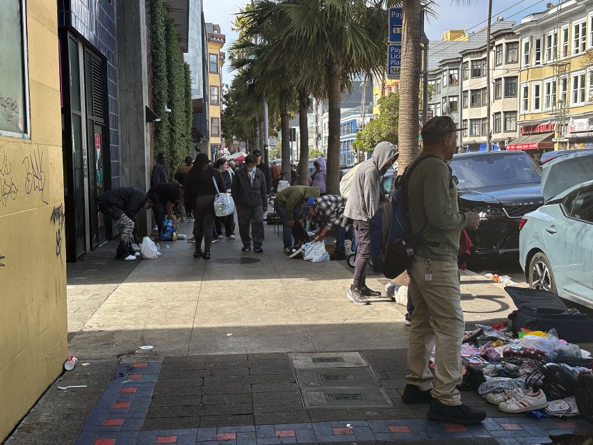 People gather on a city sidewalk among scattered belongings and bags, with parked cars and buildings in the background.
