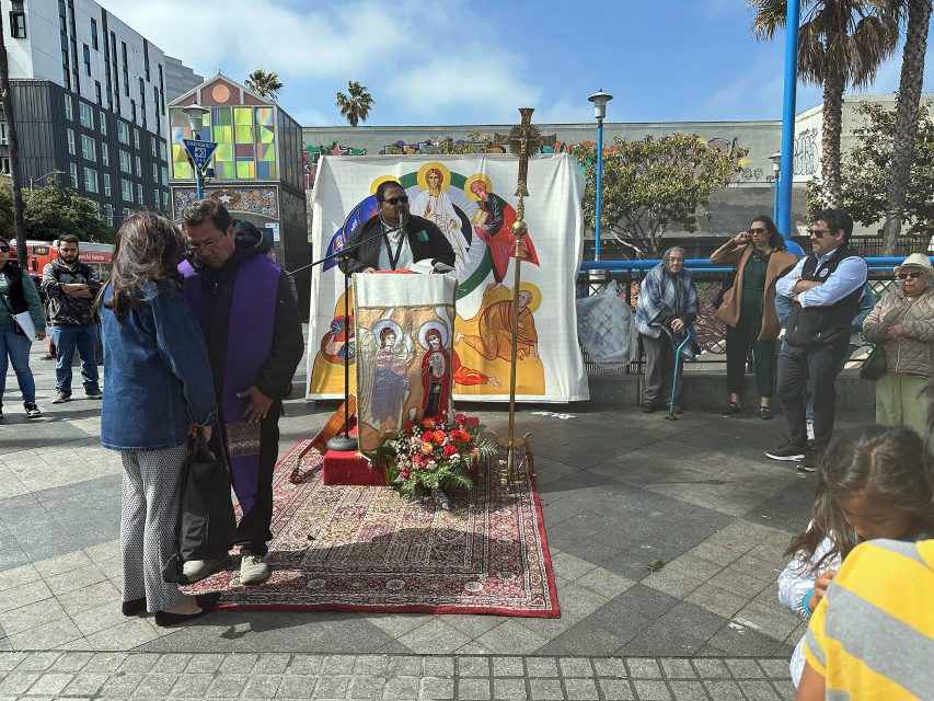 A priest speaks at an outdoor altar with religious icons, while people gather and two individuals converse in the foreground.