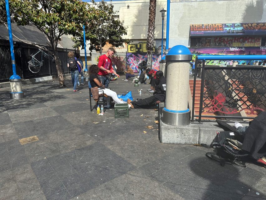 Several people are sitting and standing in an urban outdoor area with scattered belongings, graffiti-covered walls, and visible litter on the ground.