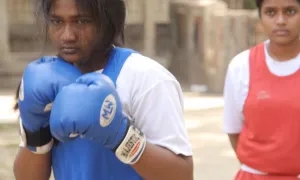 Two young women stand outdoors; the one in front wears blue boxing gloves and a blue uniform, while the one behind wears a red uniform and watches.