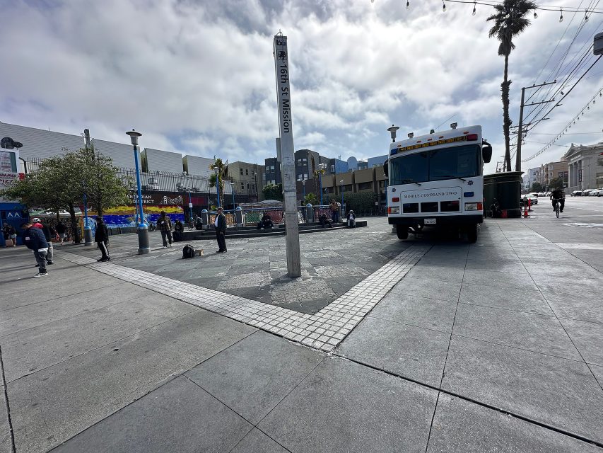 A mobile command truck is parked near an obelisk in a city plaza with people walking and sitting around on a cloudy day.