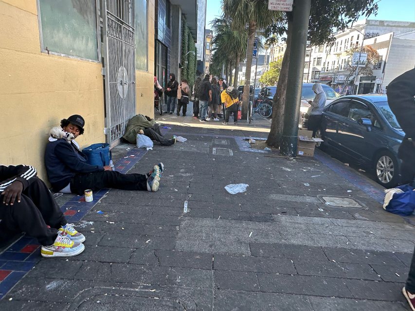 People sit and lie on a littered city sidewalk, with a group gathered in the background near parked cars and bicycles.