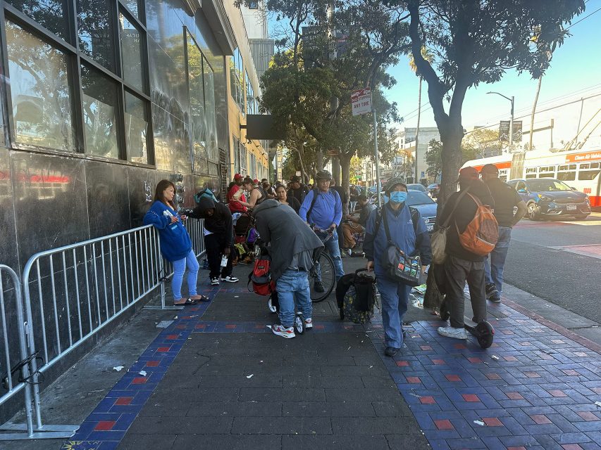 A group of people stand and sit along a city sidewalk near a building, some with bags and backpacks; trees and a bus are visible in the background.