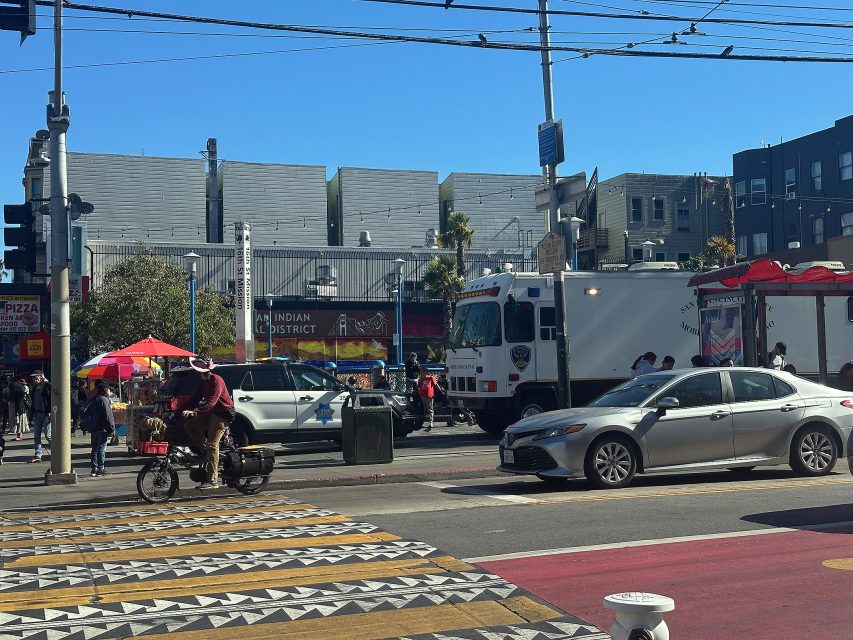 A busy urban street scene shows a man riding a bicycle cart, cars, a food truck, pedestrians, and buildings under a clear blue sky.