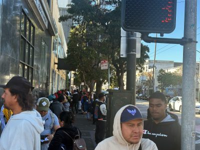 People stand in a long line on a city sidewalk near a building, with trees and street signs visible in the background.