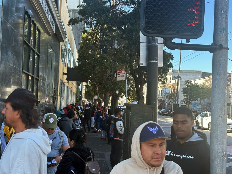 People stand in a long line on a city sidewalk near a building, with trees and street signs visible in the background.