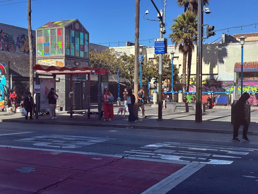 People wait at a colorful bus stop near a crosswalk; street art and murals cover surrounding walls under a clear blue sky.