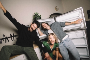 Three people pose playfully in a kitchen next to an open refrigerator, with two standing and one crouching in front of the open fridge door.