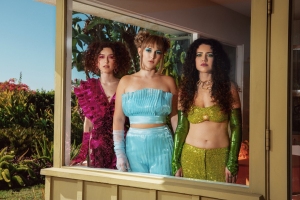 Three women in colorful, glittery outfits stand side by side and look through a window, with plants and greenery visible outside.