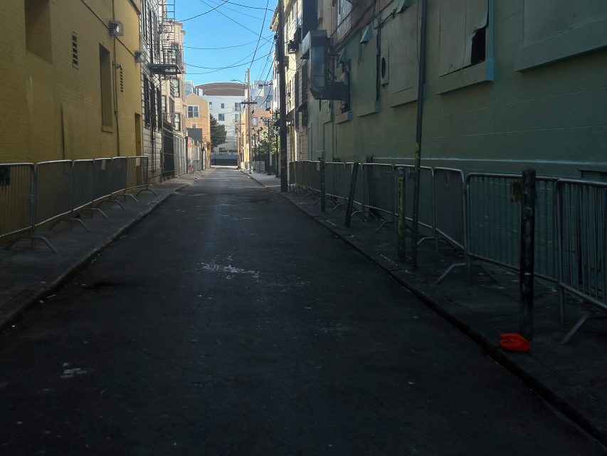 A deserted urban alleyway is bordered by metal barricades and buildings, with a red object on the ground near the right side.