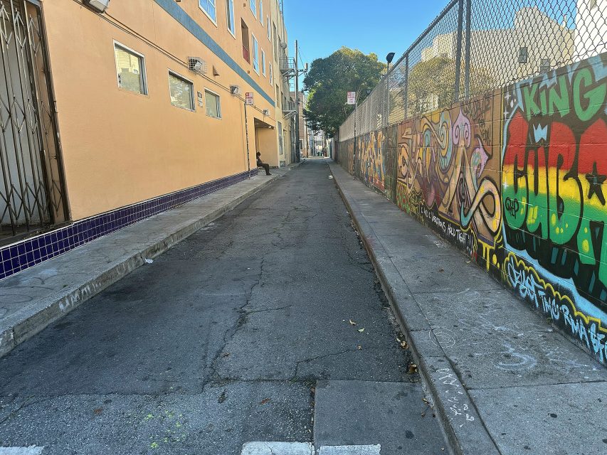 A narrow urban alleyway with graffiti on the right wall, a beige building on the left, and a cracked asphalt path running between them.