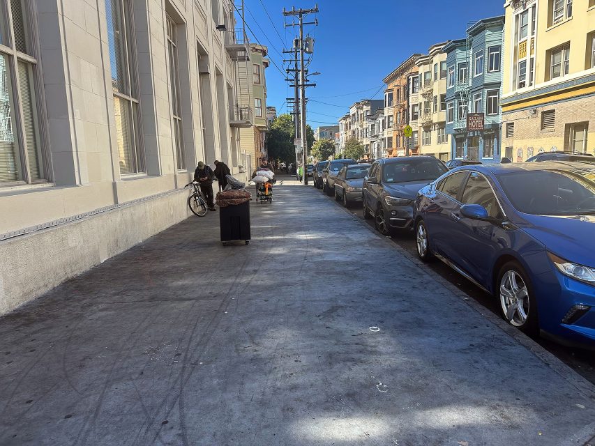 A city sidewalk with parked cars on the right, several people near a bicycle on the left, and colorful buildings lining the street under a clear blue sky.