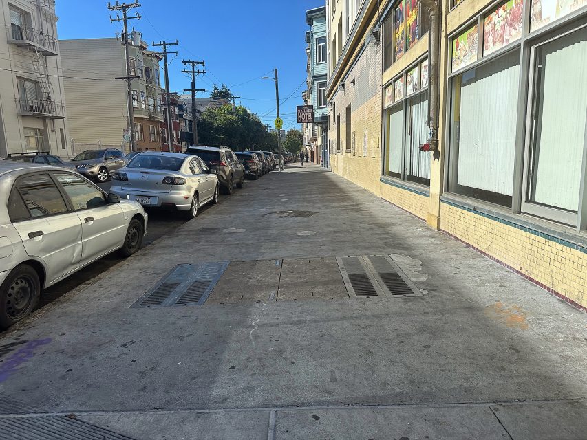 A city sidewalk with parked cars on the left and buildings with windows on the right under clear blue skies.