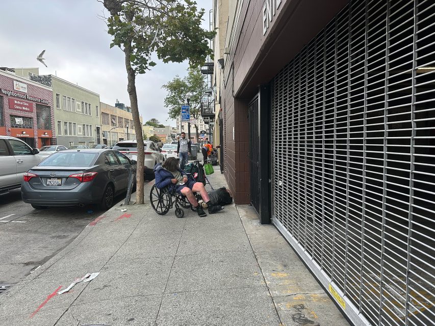 A person in a wheelchair sits on a city sidewalk beside a closed storefront, with cars parked along the street and pedestrians in the background.