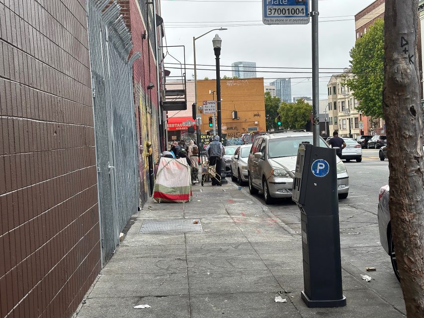 A city sidewalk with parked cars, a parking meter, people gathered near a mural wall, and buildings in the background on an overcast day.