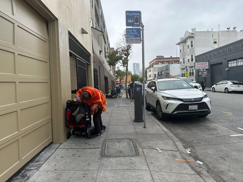 A person wearing an orange safety vest stands next to a stroller on a city sidewalk near parked cars and a pay parking sign.
