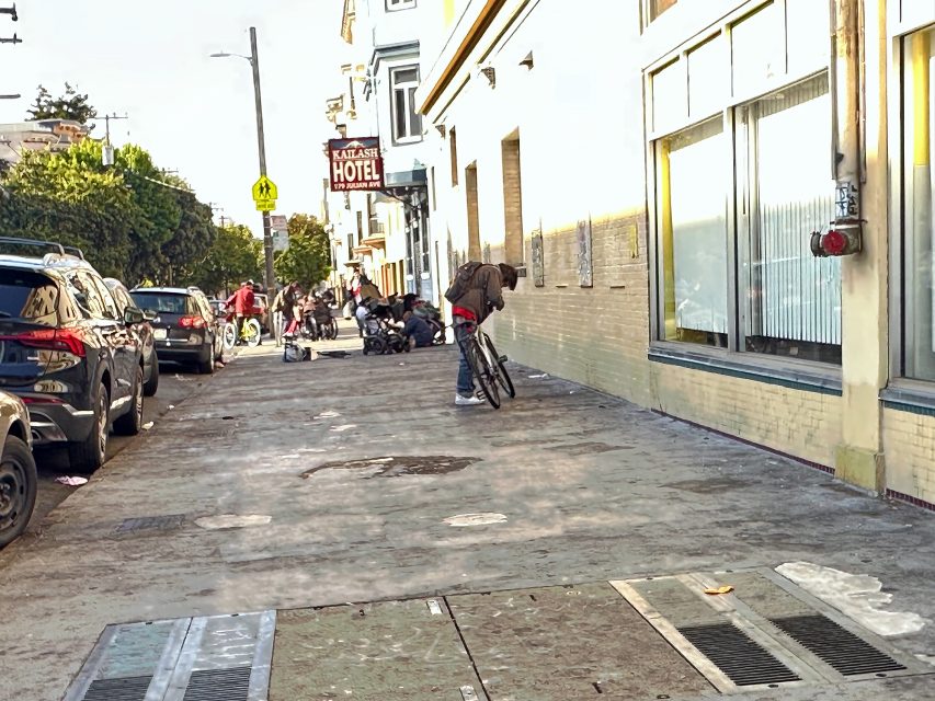 A person leans over a bicycle on a city sidewalk near parked cars, while several people and belongings are visible further down the street.