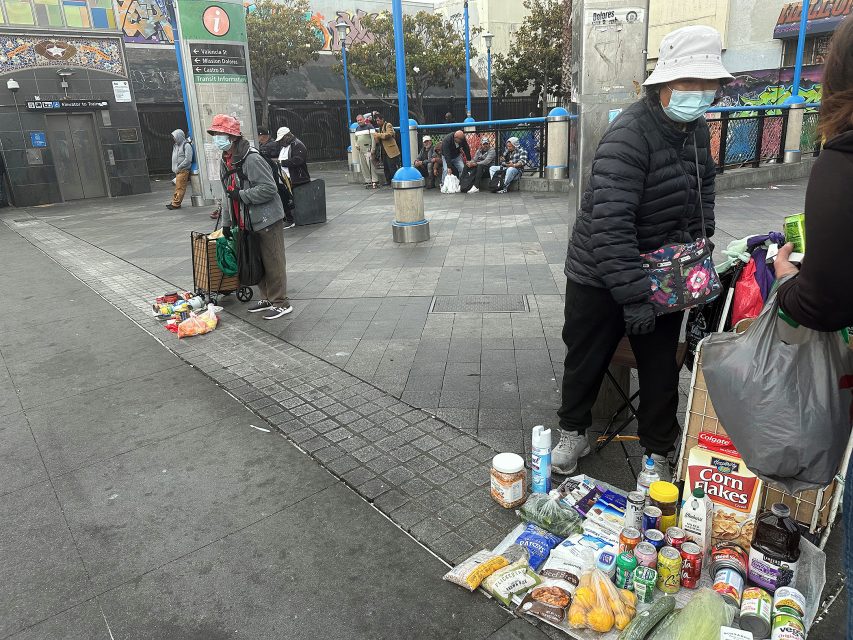 People sell various groceries and household items laid out on the pavement in an urban public square, with more people and a mural visible in the background.