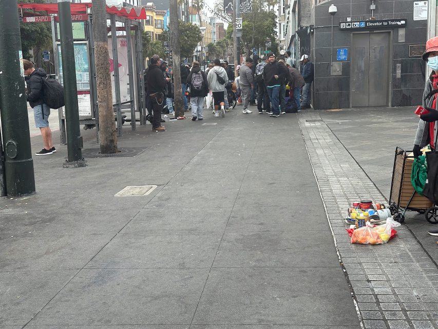 A group of people gather near a building entrance and bus stop on a city sidewalk; a cart with groceries and supplies sits in the foreground.