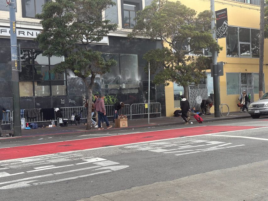 People gather and sit on a city sidewalk in front of a building with boarded-up windows and graffiti, next to trees and a red bus lane.