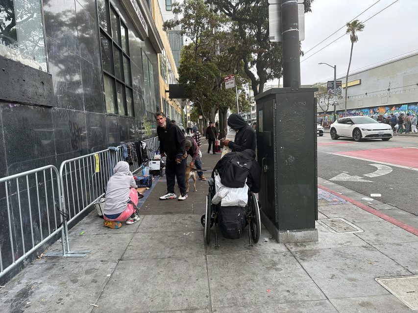 Several people with belongings and a wheelchair gather on a city sidewalk near a building and bike rack; cars and murals are visible in the background.