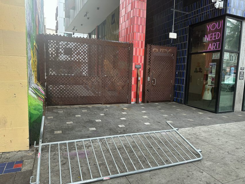 A metal barricade lies on the sidewalk in front of a gated alley next to a building with a sign that reads "YOU NEED ART.