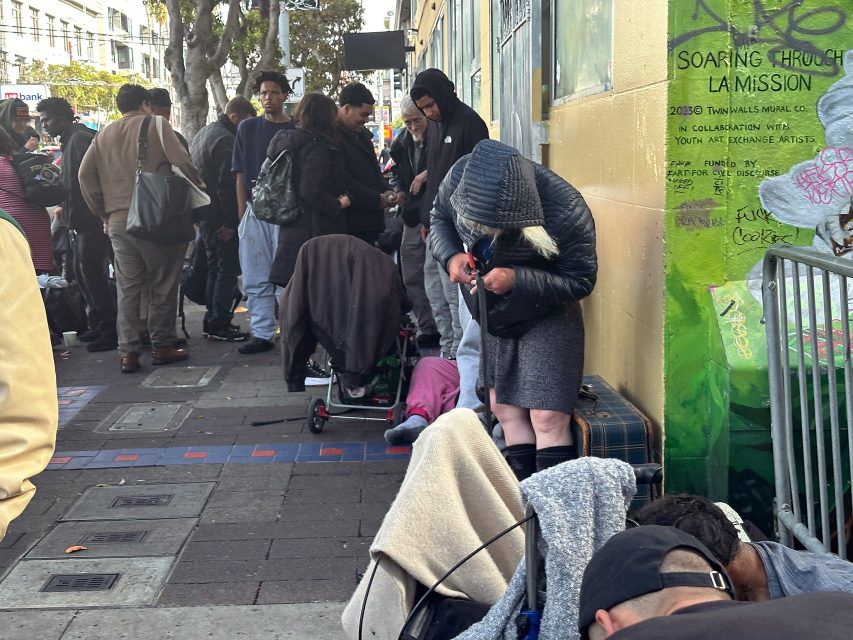 A group of people stand and sit on a city sidewalk near a building, some with belongings and carts; one woman bends over, focusing on her hands.