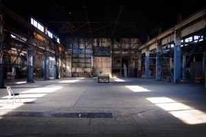 Large, empty industrial warehouse with exposed beams, high ceilings, concrete floor, one chair on the left, and a display case at the center. Sunlight streams through side windows.
