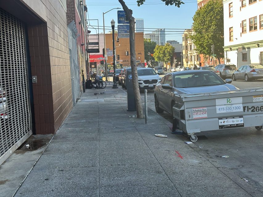 A city sidewalk with parked cars, a dumpster, and several people sitting near a building in the background on a sunny day.