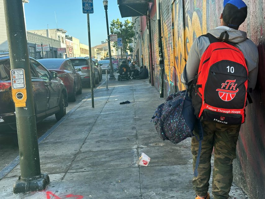 A person with a red and black backpack stands on a city sidewalk near a wall, while several people gather further down the street. Cars are parked along the curb.