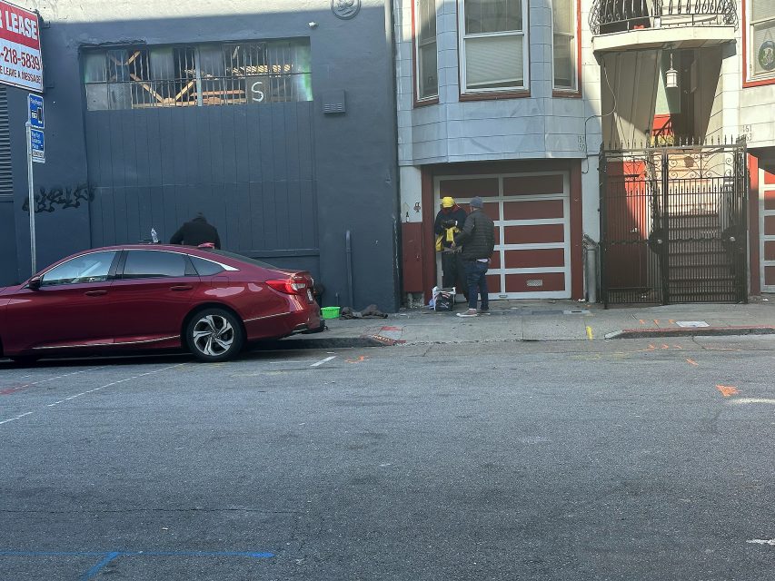 Two people stand on a sidewalk near a red car and a gray building; one wears a yellow hard hat and both appear to be working with tools or equipment at the base of a garage door.