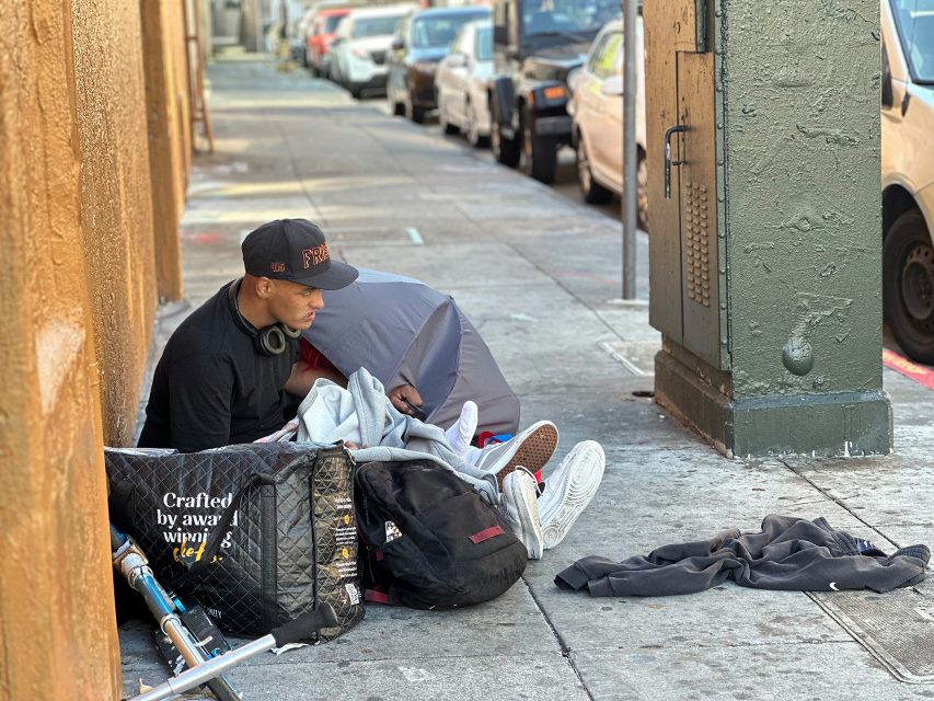 A man sits on a sidewalk surrounded by bags, clothing, and personal items, with cars parked along the street in the background.