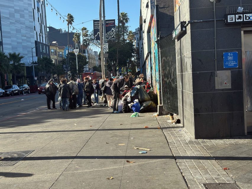 A group of people gathers on a city sidewalk next to bags of belongings, with buildings, palm trees, and parked cars visible in the background.