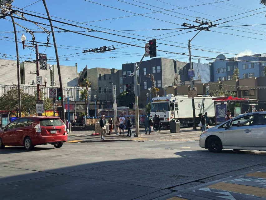 City intersection with cars stopped at a traffic light, pedestrians crossing the street, and buildings in the background under a clear sky.
