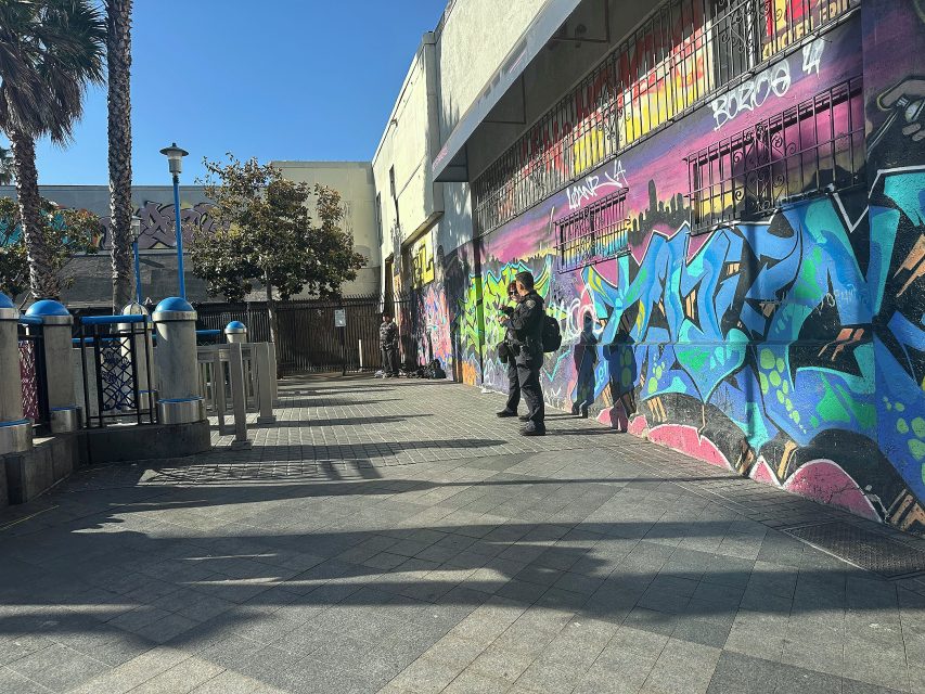 A sunlit urban sidewalk with colorful graffiti on a wall, metal fencing, palm trees, and several people walking or standing in the scene.