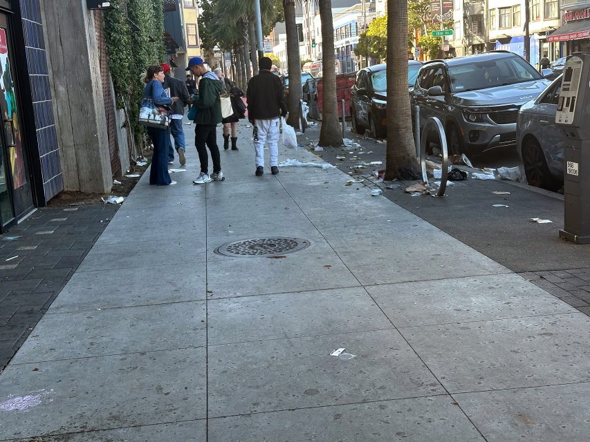 A city sidewalk with scattered trash, several parked cars, and a group of people walking and talking near buildings on a sunny day.