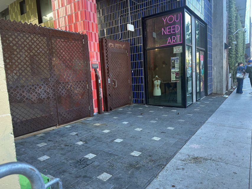 A storefront with blue and red tiled walls features a glass door and a neon sign reading "YOU NEED ART"; a fire extinguisher is mounted near a metal gate on the left.