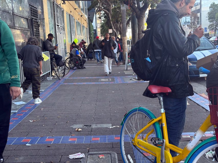 People stand and walk along a littered city sidewalk, with a yellow and teal bicycle and several pedestrians in the foreground and background.