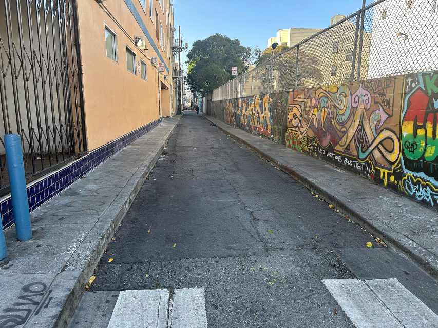 A narrow urban alleyway bordered by a building on the left and a graffiti-covered fence on the right, with trees and more buildings visible in the distance.