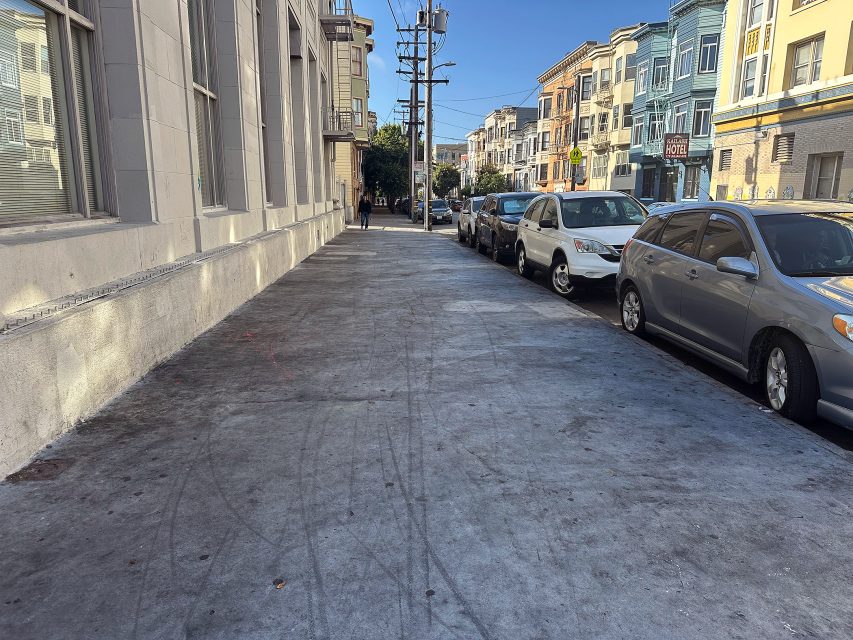A city sidewalk runs alongside parked cars and multi-story buildings on a sunny day. Two people are visible in the distance near the end of the block.