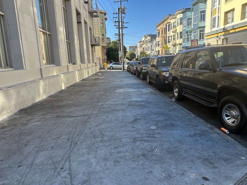 A city sidewalk next to parked cars and colorful buildings on a clear day, with some scattered litter on the ground.