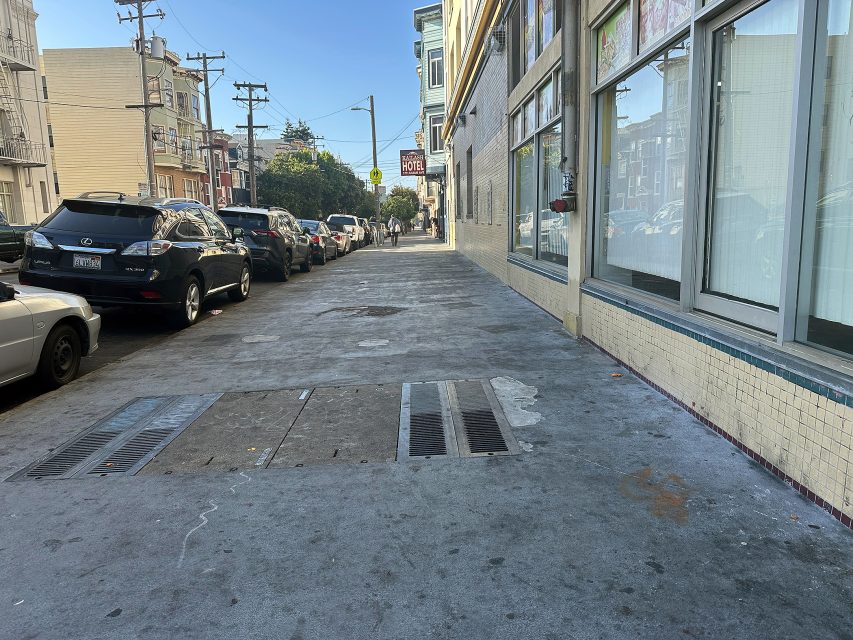 A city sidewalk runs alongside parked cars and a row of buildings under a clear blue sky.