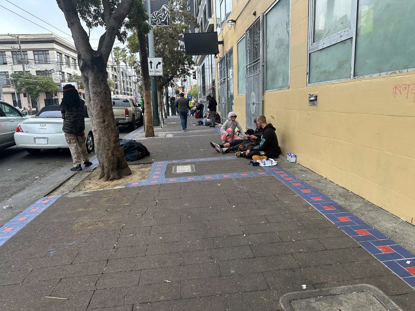 Several people sit on a sidewalk against a building, surrounded by belongings, while others walk or stand nearby on an urban street.