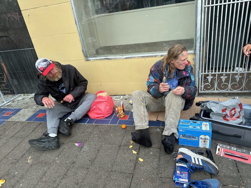 Two people sit on the sidewalk with belongings and snacks scattered around them, next to a building with a barred door and window.