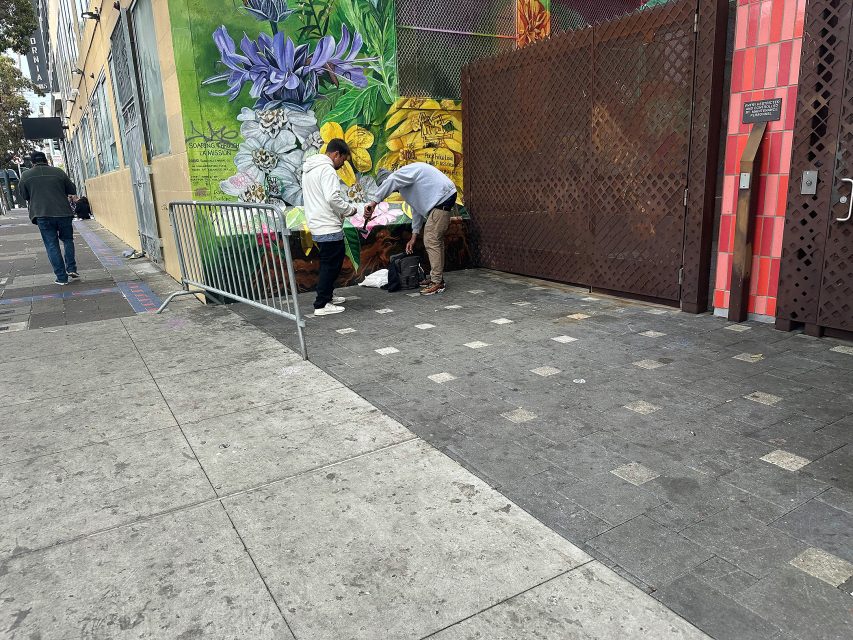 Two people stand near bags and belongings by a mural-covered wall and large gate on a city sidewalk, with a metal barricade nearby.