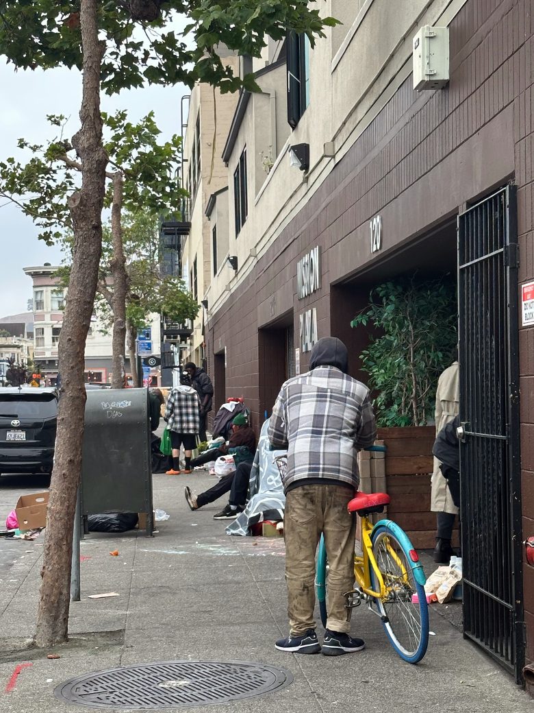 A person with a plaid jacket stands next to a bicycle on a city sidewalk, with several others and scattered belongings nearby in front of a building.