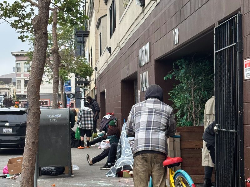 A person with a plaid jacket stands next to a bicycle on a city sidewalk, with several others and scattered belongings nearby in front of a building.